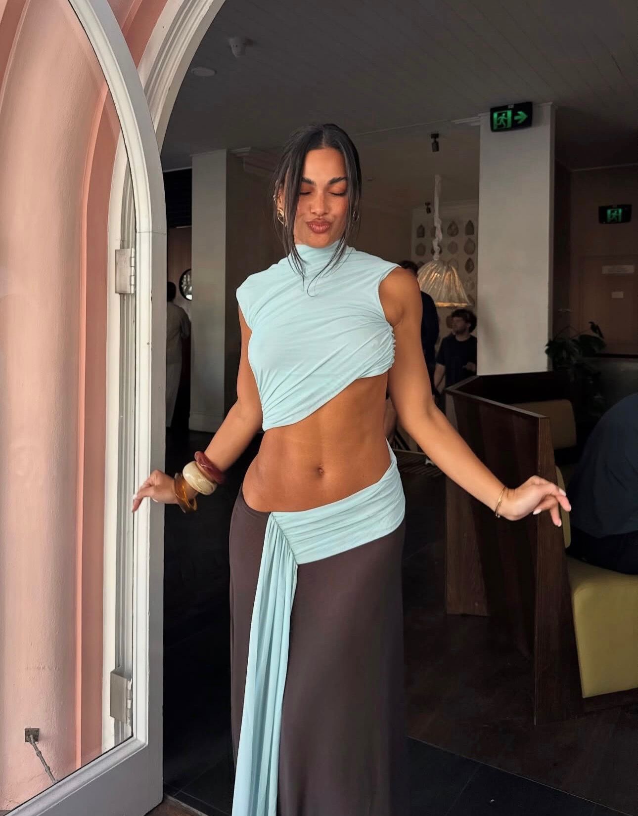 A woman posing in a light blue asymmetrical wrap top and a matching dark brown wrap skirt inside a cafe or restaurant setting.
