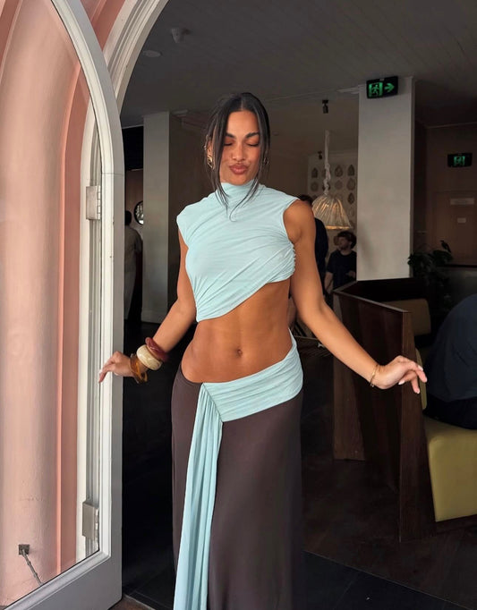 A woman posing in a light blue asymmetrical wrap top and a matching dark brown wrap skirt inside a cafe or restaurant setting.