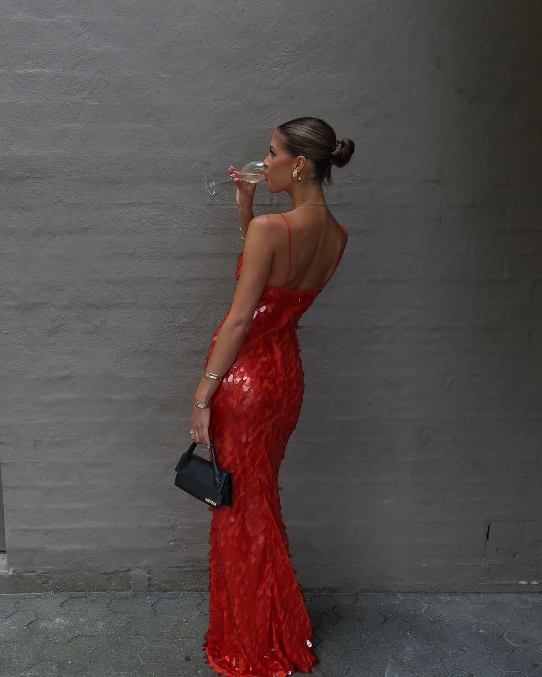 A woman in a shimmering red sequined mermaid gown drinks from a glass while holding a small black clutch against a gray textured wall.