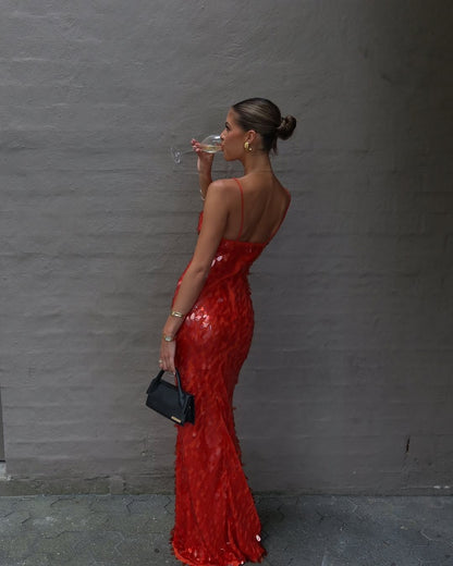 A woman in a shimmering red sequined mermaid gown drinks from a glass while holding a small black clutch against a gray textured wall.