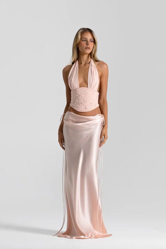 A woman modeling a pale blush pink two-piece outfit consisting of a halter crop top with lace applique and a matching satin wrap skirt, shot against a light gray studio background.