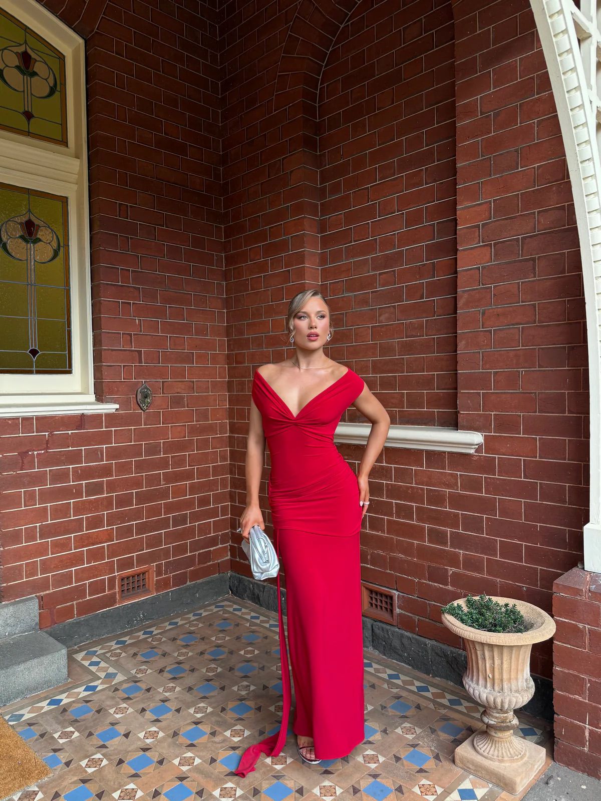 Woman in a red dress standing in front of a brick wall with a decorative plant and window.