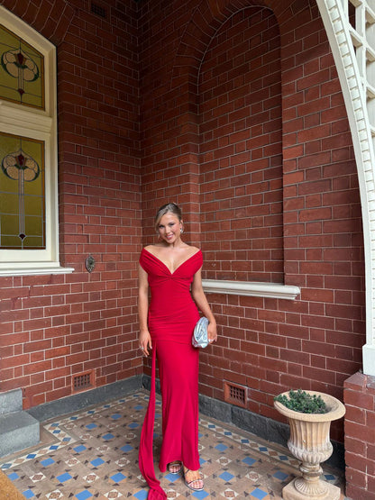 A woman wearing a vibrant red off-the-shoulder evening gown with a thigh-high slit, holding a small silver clutch, standing on a brick porch with patterned tile flooring.