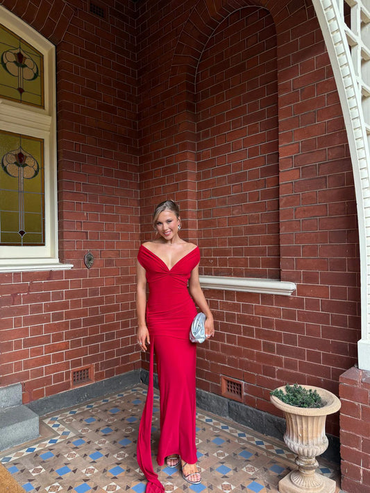 A woman wearing a vibrant red off-the-shoulder evening gown with a thigh-high slit, holding a small silver clutch, standing on a brick porch with patterned tile flooring.