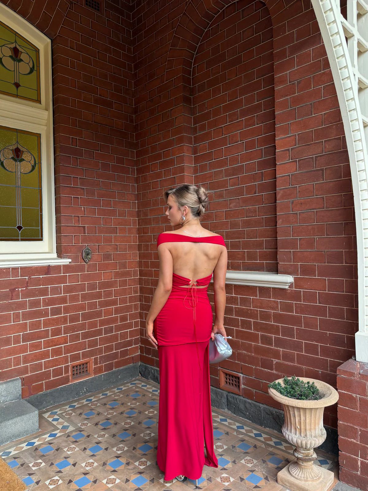 A woman wearing a bright red off-the-shoulder maxi dress with an open back and tie detail, standing in a brick building corner with patterned tile flooring.