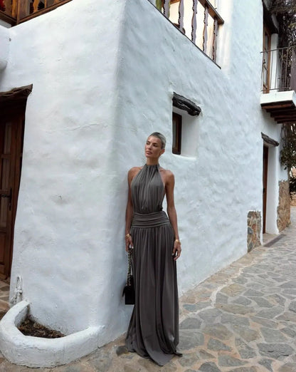 A woman wearing a dark gray halter-neck maxi dress stands in front of a white stucco building with a stone-paved walkway.