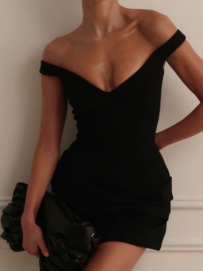 A woman wearing a fitted off-the-shoulder black mini dress, holding a black ruffled clutch against a light beige wall.