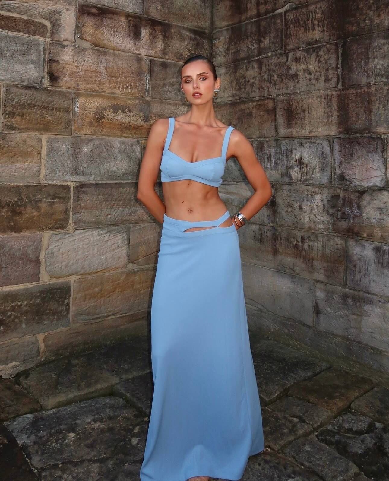 A woman stands in a stone-walled corner wearing a light blue two-piece outfit consisting of a cropped bandeau-style top and a long skirt with a side cutout.