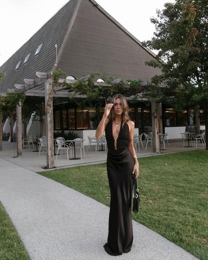 A woman stands on a paved walkway in front of a modern building with a large angled roof, grass, and trees. She is wearing a floor-length black satin-like gown with a plunging neckline and carries a small black handbag.