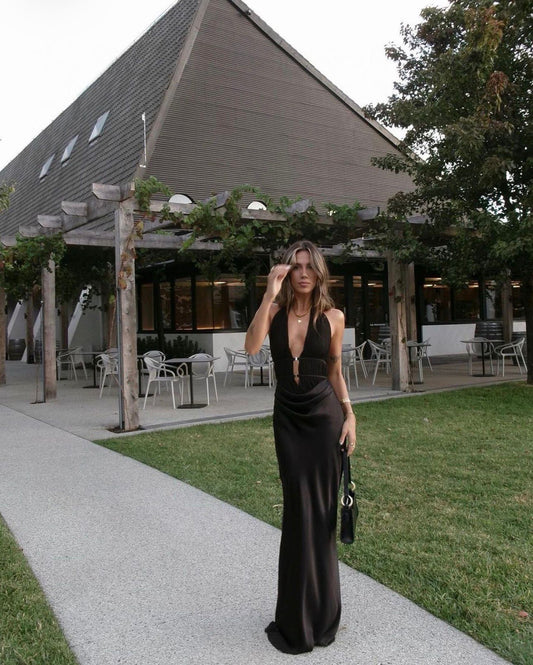 A woman stands on a paved walkway in front of a modern building with a large angled roof, grass, and trees. She is wearing a floor-length black satin-like gown with a plunging neckline and carries a small black handbag.