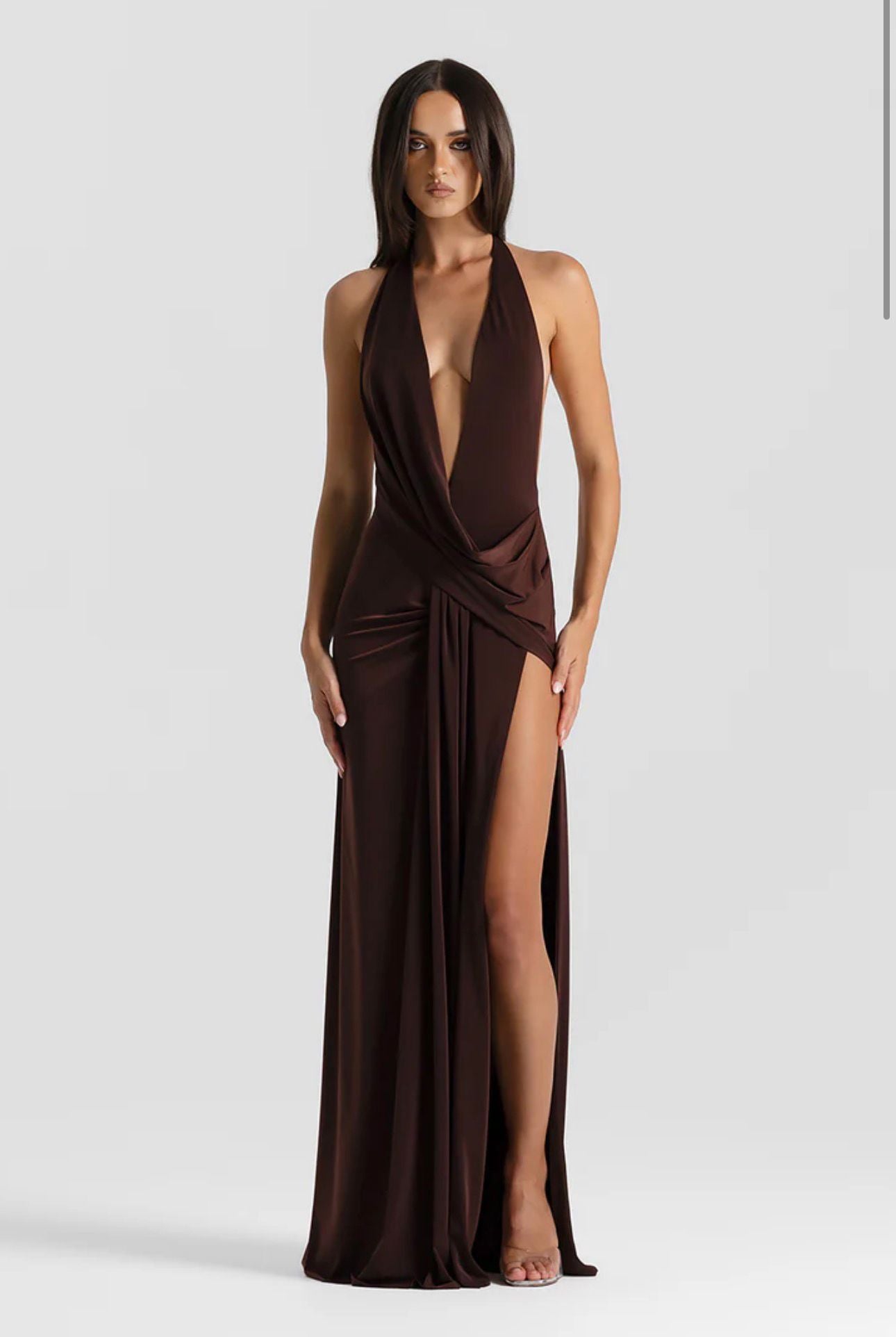 A woman modeling a floor-length dark brown halter gown with a deep plunging V-neckline, draped wrap-style bodice, and a high thigh-high slit, photographed against a light gray studio background.