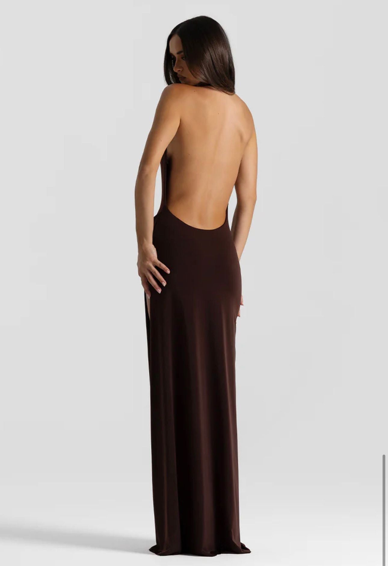 A woman modeling a floor-length, open-back gown from the back against a light gray studio background. The dress is a solid dark brown color with a smooth, satin-like texture.
