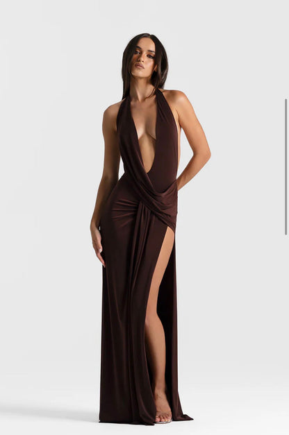 A woman modeling a floor-length dark brown halter gown with a plunging V-neckline, draped waist detail, and a high thigh slit, photographed against a light gray studio background.