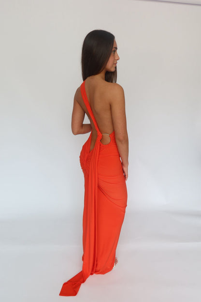 A woman modeling a bright orange-red draped maxi dress from the back, featuring a single strap and a long train, set against a plain white studio background.