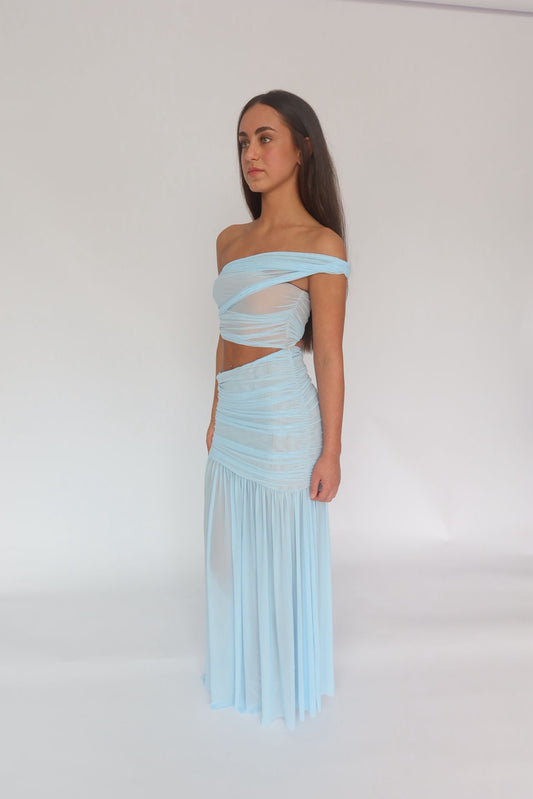 A woman modeling a light blue off-the-shoulder two-piece set with a ruched cutout top and a matching pleated maxi skirt, photographed against a plain white studio background.