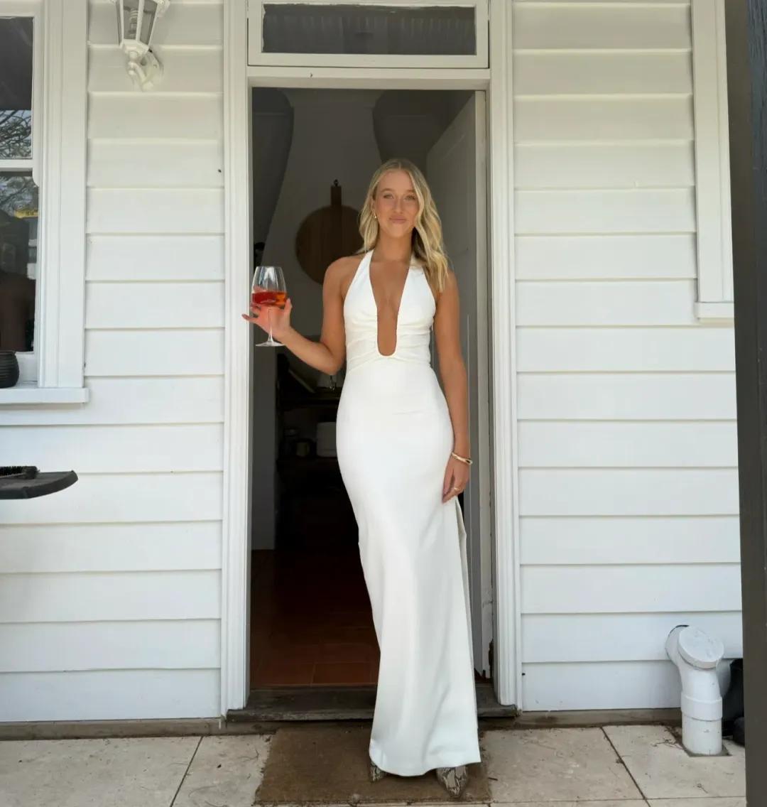 A woman stands in a doorway wearing a fitted white sleeveless gown with a deep plunging neckline and a floor-length silhouette. She holds a wine glass in one hand, with a white house exterior behind her.