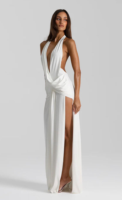 A woman modeling a floor-length white halter gown with a deep plunging V-neckline, draped waist detail, and a high thigh-high slit, photographed against a light gray studio background.