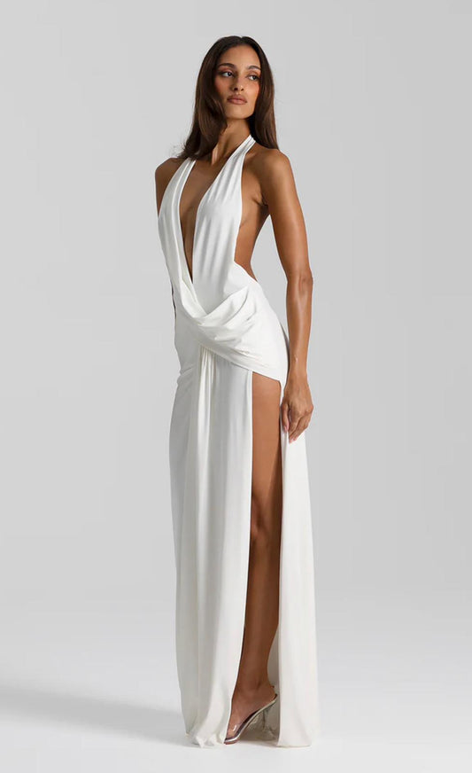 A woman modeling a floor-length white halter gown with a deep plunging V-neckline, draped waist detail, and a high thigh-high slit, photographed against a light gray studio background.