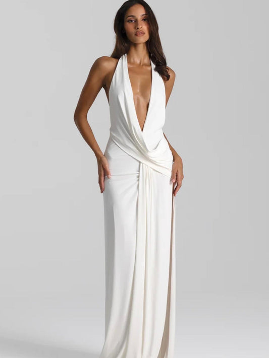 A woman modeling a floor-length white halter gown with a plunging V-neckline and draped wrap detail at the waist, photographed against a light gray studio background.