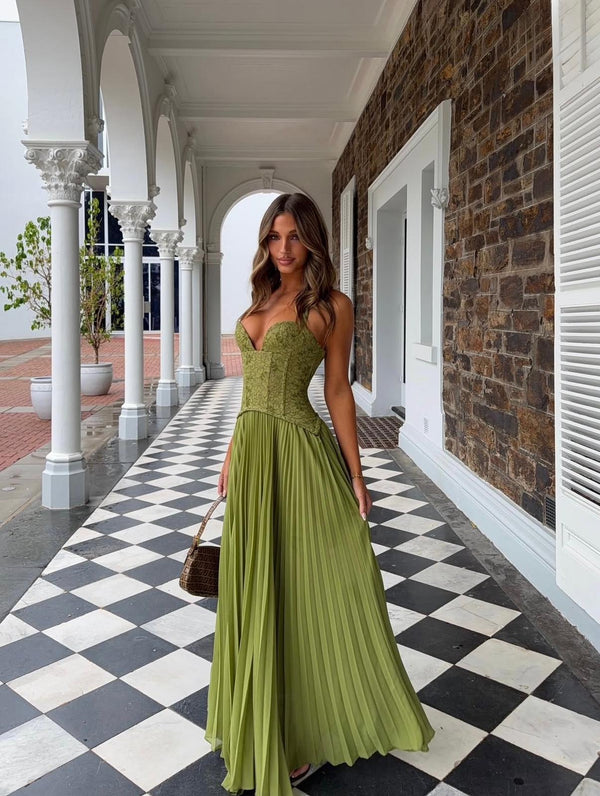 A woman wearing a sleeveless olive green formal gown with a structured bodice and flowing pleated skirt, standing in a covered walkway with a black-and-white checkered floor.