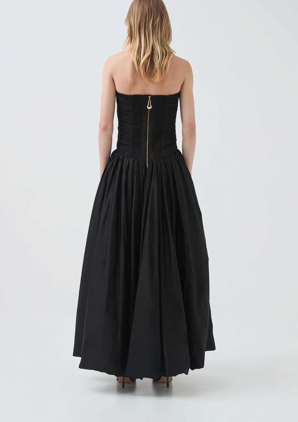 A woman viewed from the back wearing a strapless black gown with a structured corset-style bodice, full gathered skirt, and a visible gold-toned center back zipper. The photo is taken in a clean studio setting with a light gray background.