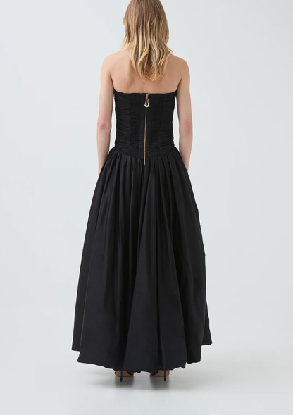 A woman viewed from the back wearing a strapless black gown with a structured corset-style bodice, full gathered skirt, and a visible gold-toned center back zipper. The photo is taken in a clean studio setting with a light gray background.